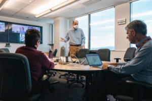 Office collaboration session during hybrid team meeting with workers seating around round table engaged in discussion with virtual attendees on monitor screen