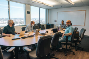 Office collaboration session during hybrid team meeting with workers seating around round table engaged in discussion with virtual attendees on monitor screen