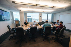 Office collaboration session during hybrid team meeting with workers seating around round table engaged in discussion with virtual attendees on monitor screen