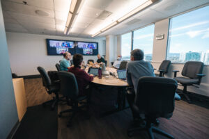 Office collaboration session during hybrid team meeting with workers seating around round table engaged in discussion with virtual attendees on monitor screen